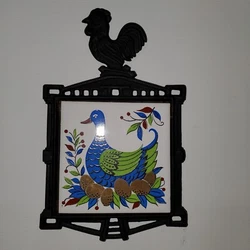 Cast iron trivet with the handle in the shape of a rooster.  Ceramic tile has blue and green goose sitting on nest of...