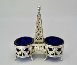 The double salt cellar in silver and blue glass resting on shoe feet, the openwork body decorated with putti, garlands...