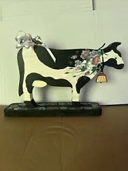 Vintage Wooden Cow With Painted Decorative Flowers On The Cow And Stand. The cow has a bell around it’s neck and...